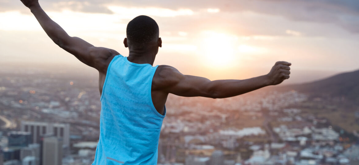 Carefree independent man with dark skin, stands back at camera, stretches hands as holding world, feels freedom, wears blue casual vest, enjoys panoramic view with sunrise. Recreation concept