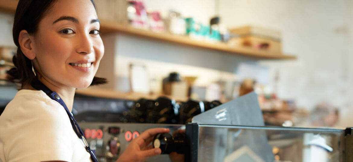 Vertical shot of barista, asian girl steaming milk for cappuccino, prepare latte for client, wearing blue apron, smiling happily, working in cafe