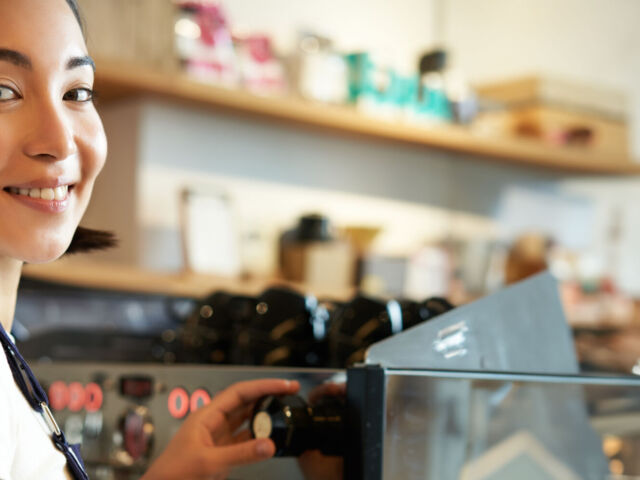 Vertical shot of barista, asian girl steaming milk for cappuccino, prepare latte for client, wearing blue apron, smiling happily, working in cafe