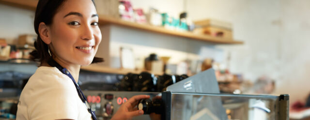 Vertical shot of barista, asian girl steaming milk for cappuccino, prepare latte for client, wearing blue apron, smiling happily, working in cafe